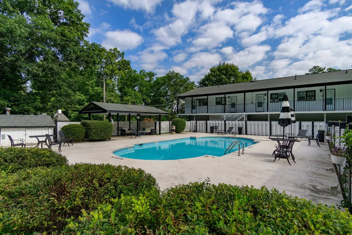 The image showcases the inviting exterior of the Mill House Nest condo, with a sparkling swimming pool at the center, surrounded by lush greenery. Comfortable outdoor seating areas and a charming gazebo promise both relaxation and opportunities for socializing. The tranquil atmosphere is enhanced by the late afternoon or sunset light, hinting at leisurely swims, shaded lounging, and memorable evenings spent outdoors.