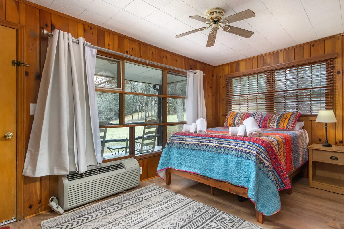 This interior photo features a queen-sized bed with a colorful bedspread, a large window letting in abundant natural light, wood paneled walls, a decorative rug, and a nightstand with a softly glowing lamp. The scene exudes warmth and tranquility, offering a glimpse of a restful sleeping space that encourages relaxation and comfort.