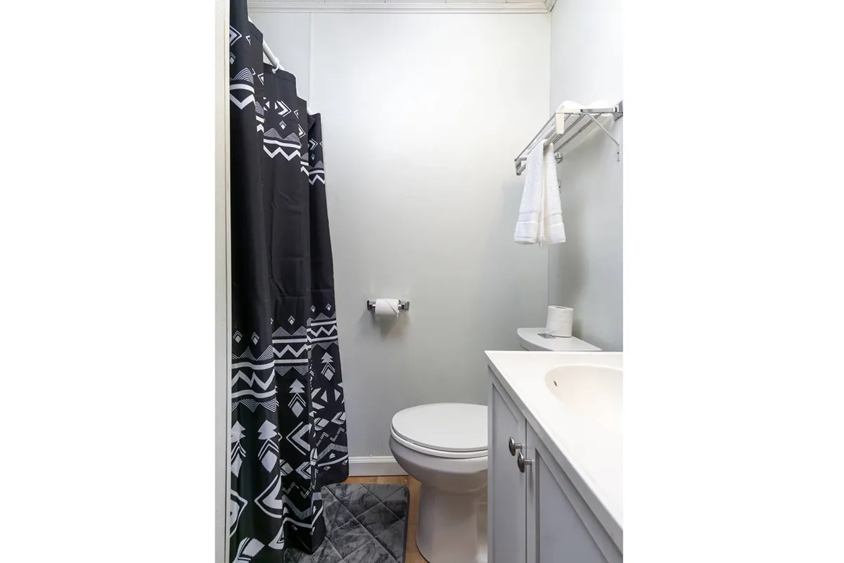 The ensuite bathroom is presented with a minimalist aesthetic, featuring a white vanity, a toilet, and a shower with a geometric-patterned curtain. The space is clean, modern, and private, offering a refreshing and functional setting for self-care and relaxation.