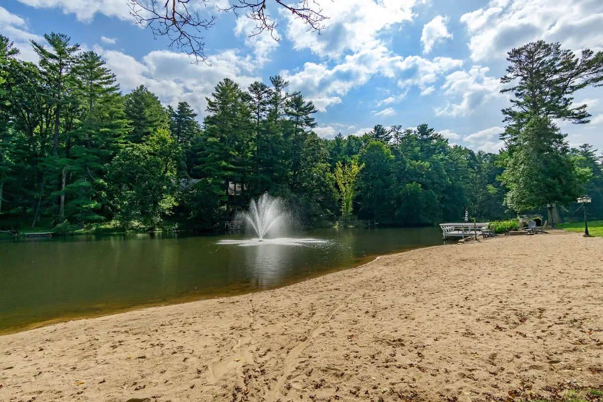 A tranquil lakeside scene featuring a sandy beach, a fountain in the water, a dock, and surrounding trees. The area looks inviting for swimming, sunbathing, fishing, picnicking, and boating, with the gentle curve of the beach and calm water creating a relaxing resort atmosphere.