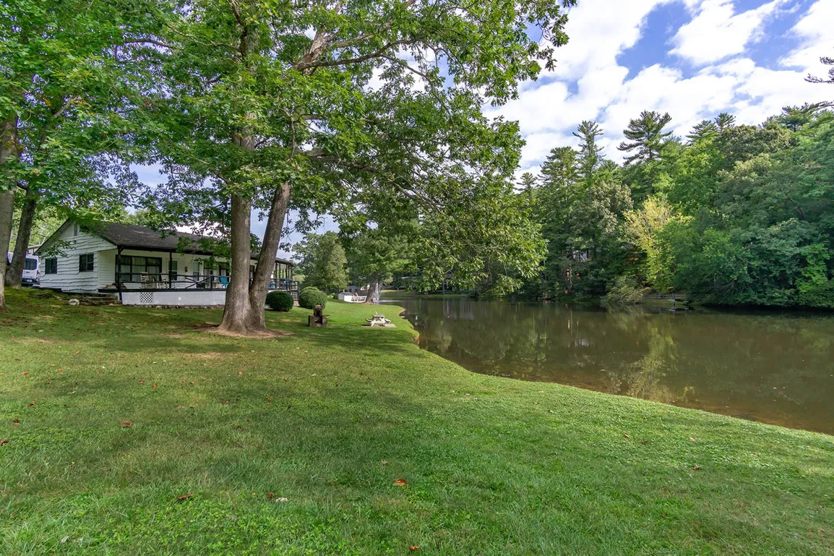 This image features the house set against a tranquil lake, bordered by mature trees and an open lawn. The scene conveys an inviting, peaceful environment ideal for leisure activities like fishing, strolling, and relaxing by the water—perfect for guests accompanied by their pets.