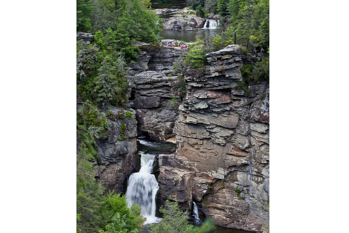 A scenic view of waterfalls cascading over rock formations, surrounded by lush greenery and people enjoying the outdoors. The image conveys the nearby hiking and nature experiences available in the Blue Ridge region.
