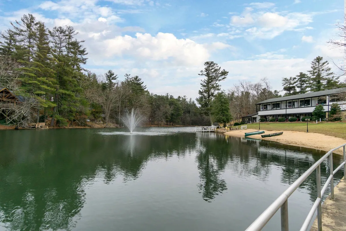A tranquil view featuring a lake with a central fountain, surrounded by lush trees, a sandy beach area, and the facade of a building in the background. The beach and grass invite families to play, picnic, or relax by the water, evoking the essence of lakeside fun and outdoor exploration.