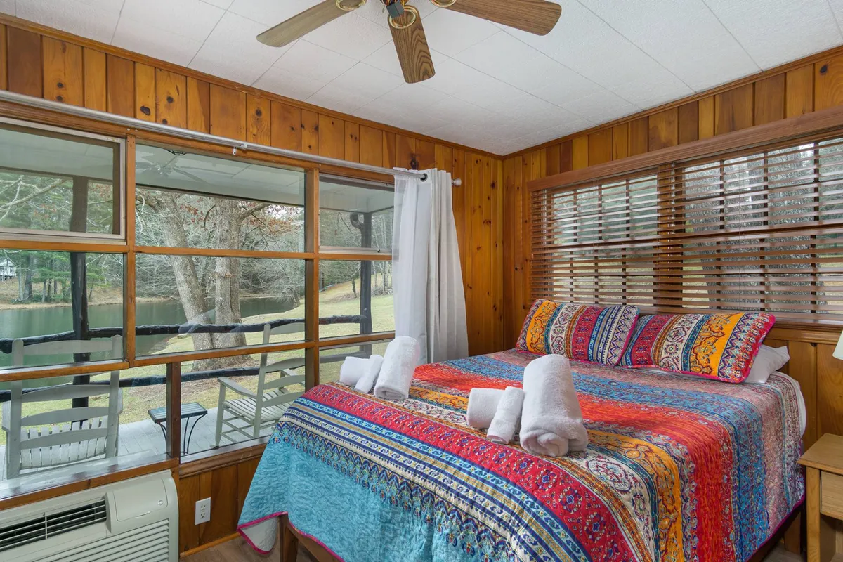 A tranquil bedroom scene showcasing a colorful bedspread, large windows overlooking nature, and wood paneling that adds to the cozy, cabin-like ambiance. The space is bathed in natural light, emphasizing the relaxing environment and connection to the outdoors.