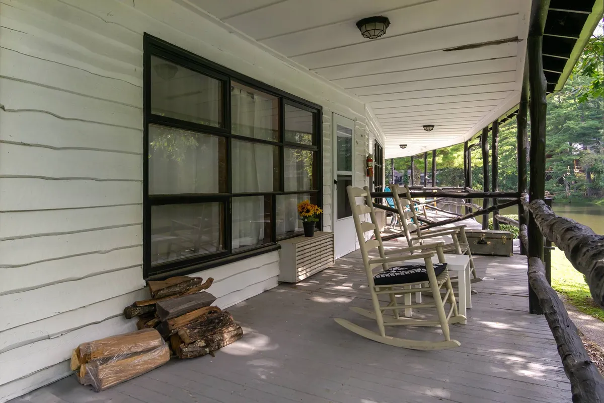 A cozy porch scene with two rocking chairs, a neat woodpile, a window adorned with flowers, and views of surrounding nature. The tranquil setting is ideal for relaxing mornings or evenings in the fresh air with pets nearby.