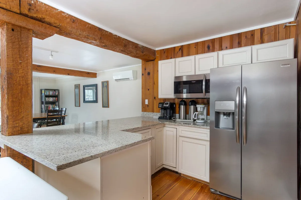 A spacious, modern kitchen with granite countertops, stainless steel refrigerator, warm wooden cabinetry, and exposed wooden beams overhead. The area features ample prep space and is designed for both cooking and entertaining, with a welcoming, homey ambiance.
