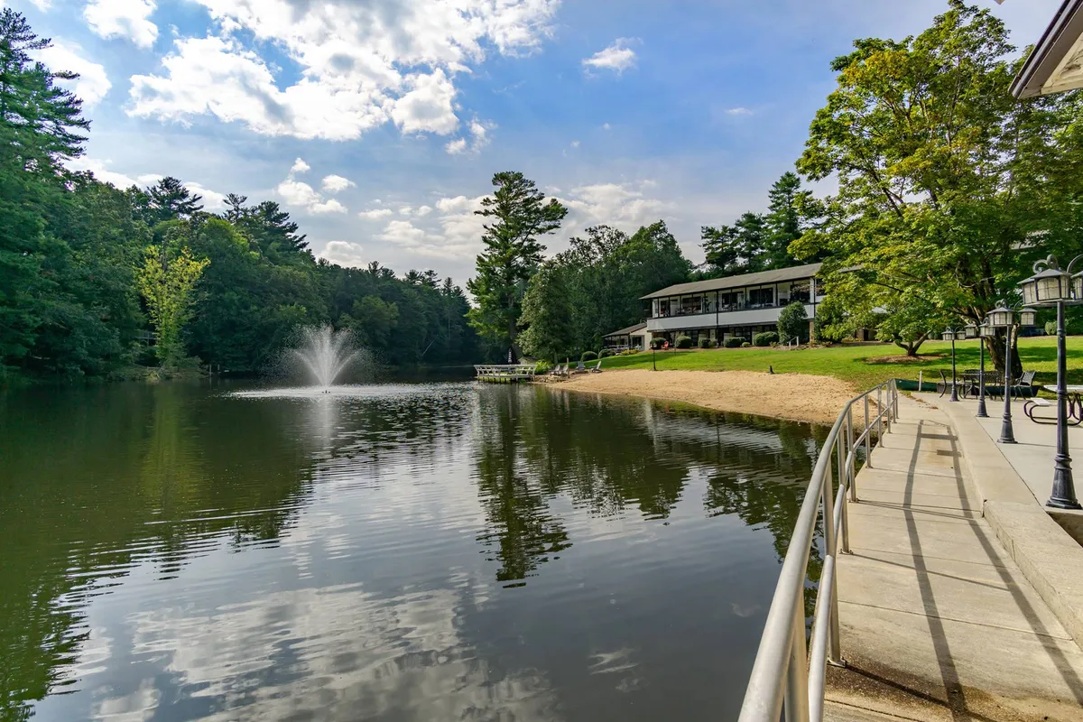 This image displays a tranquil lake with a calm surface, a fountain, a sandy beach area, surrounding trees, and the property building in the background. The scene is serene and inviting, suggesting opportunities for relaxation, boating, and enjoying nature.