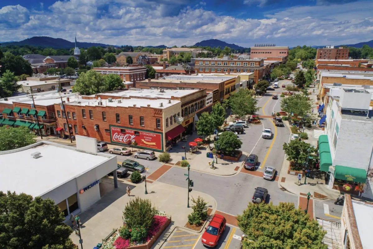 A lively streetscape view of Flat Rock Village features charming historic buildings, tree-lined streets, and pedestrians enjoying the local scene. The Coca-Cola advertisement and street life reinforce the vibrancy and small-town appeal, inviting guests to explore eateries, shops, and cultural attractions.