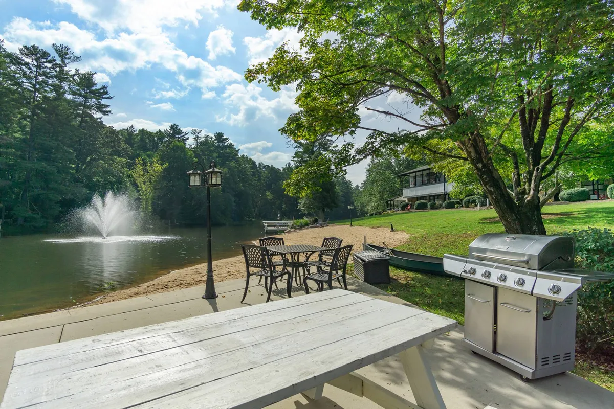A lively lakeside setting with a grill, picnic table, fountain, chairs, and the lake in the background. The scene suggests a location where families gather for barbecues, picnics, fishing, and relaxing by the water—ideal for group activities and memory-making.