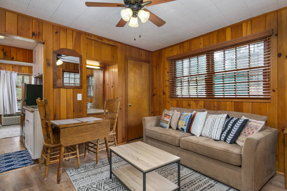 A warm, inviting living and dining area featuring a plush sofa with decorative pillows, a textured area rug, and a wooden dining table with chairs. Natural light streams through large windows, creating a bright, welcoming environment. The space showcases rustic-boho decor elements and a cozy layout ideal for both relaxation and dining.