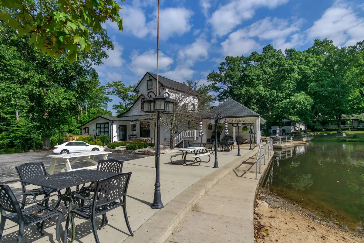 This image captures the main building of the resort with an inviting outdoor seating area set beside a picturesque lake and surrounded by mature trees. The tranquil waters and communal seating evoke opportunities for dining al fresco, sharing local wines, or simply relaxing in a cozy outdoor environment. The image radiates a sense of seclusion and exclusivity.